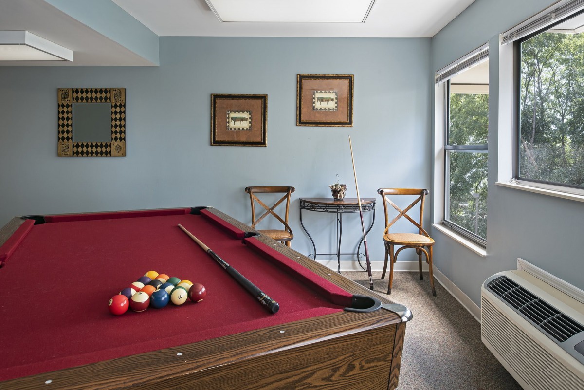 A cozy game room featuring a red-felt pool table, accompanied by chairs and a small table beside a bright window.