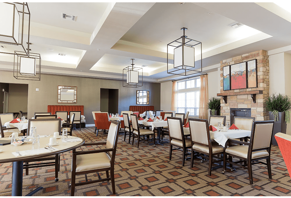 A well-lit dining room with neatly arranged tables and chairs, featuring a patterned carpet and modern lighting fixtures.