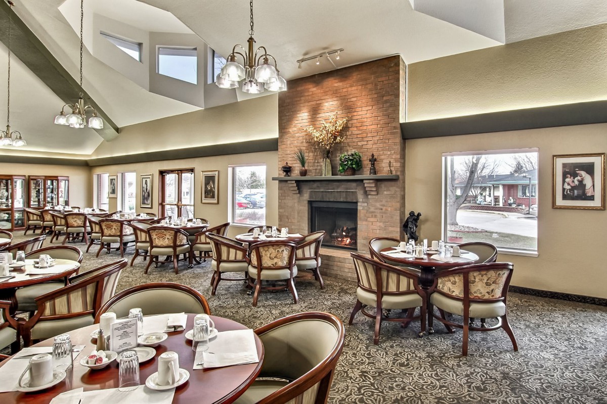 A cozy restaurant dining area featuring round tables, elegant chairs, and a warm fireplace surrounded by large windows.
