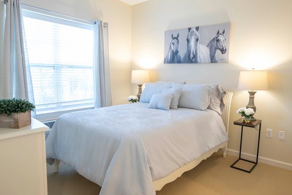 A cozy bedroom with a neatly arranged bed, soft lighting, and a painting of horses on the wall.