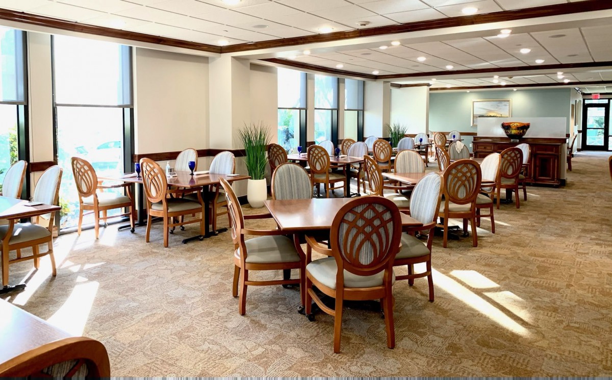 An empty dining room with several wooden tables and chairs, featuring large windows and a carpeted floor.