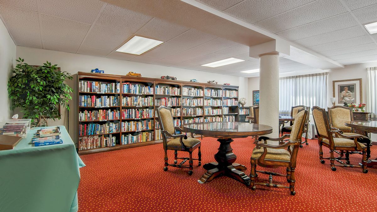 A cozy library room with bookshelves filled with books, a round table, and comfortable chairs on a red carpeted floor.