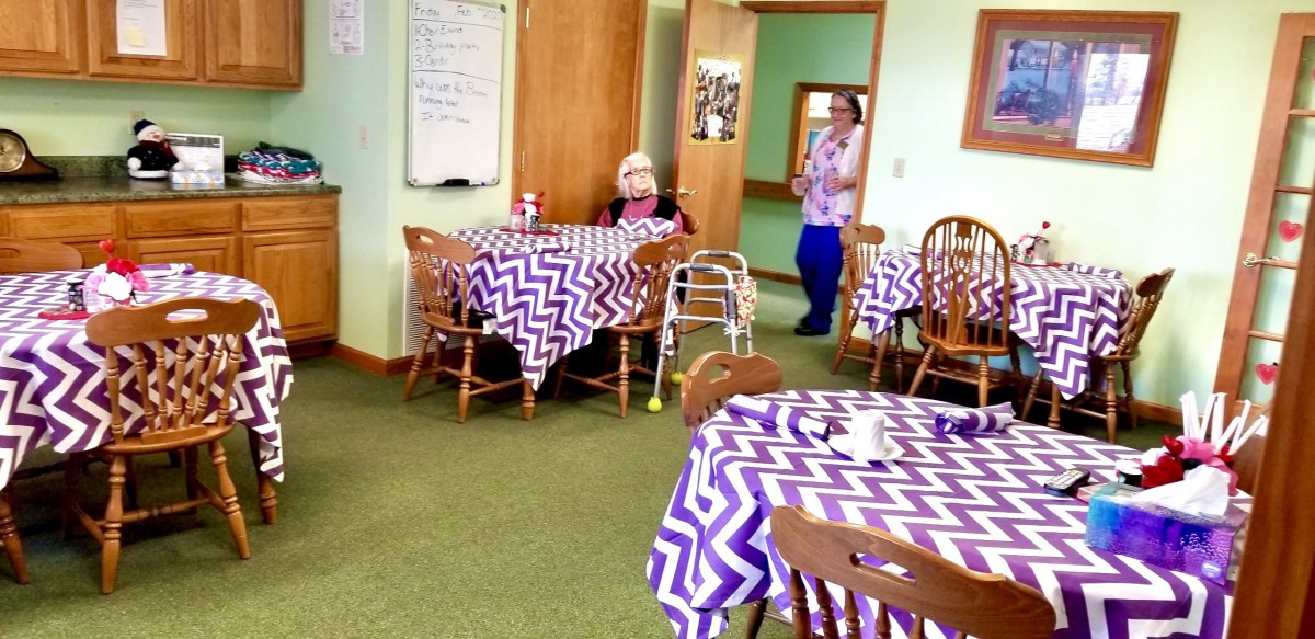 An elderly person sits in a dining room with zigzag-patterned tablecloths, accompanied by a caregiver.