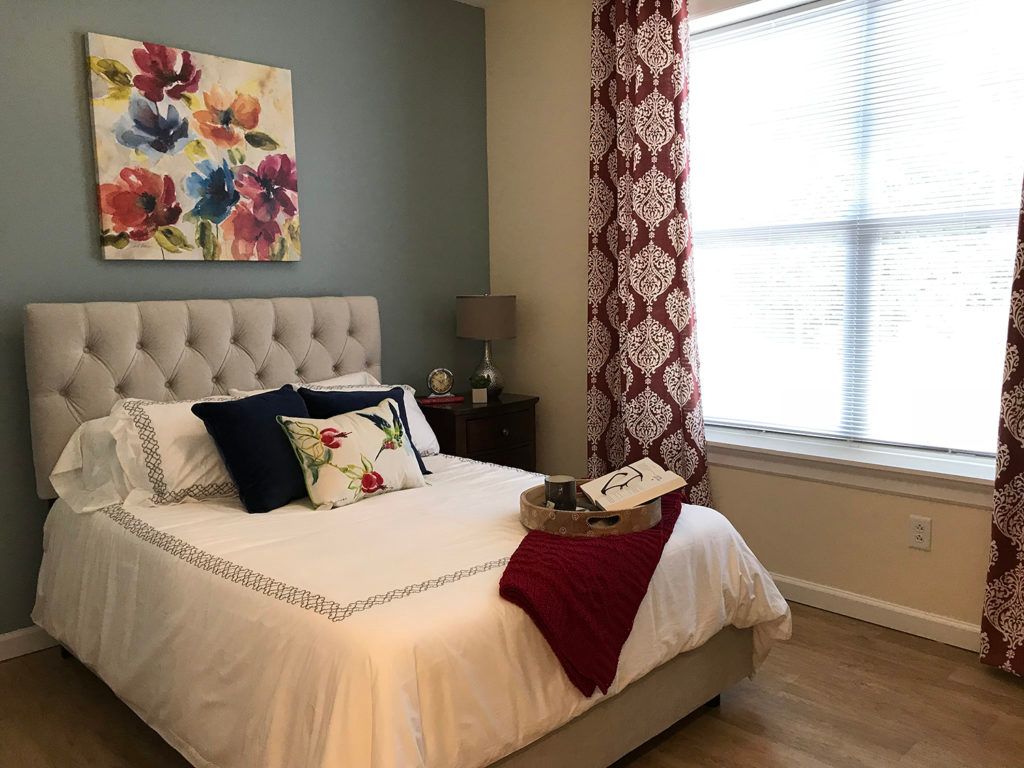 A cozy bedroom with a neatly made bed, featuring floral artwork and decorative pillows, complemented by patterned curtains and a nightstand with a lamp.