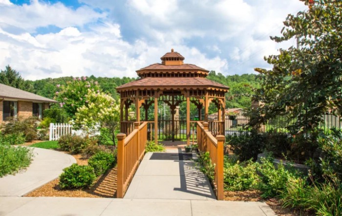 A scenic view of a wooden gazebo connected by a bridge in a lush garden setting.
