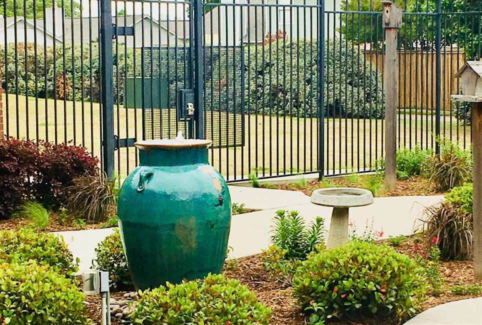 A tranquil garden scene featuring a large green vase and a birdbath amidst neatly trimmed shrubs, with a tall fence in the background.