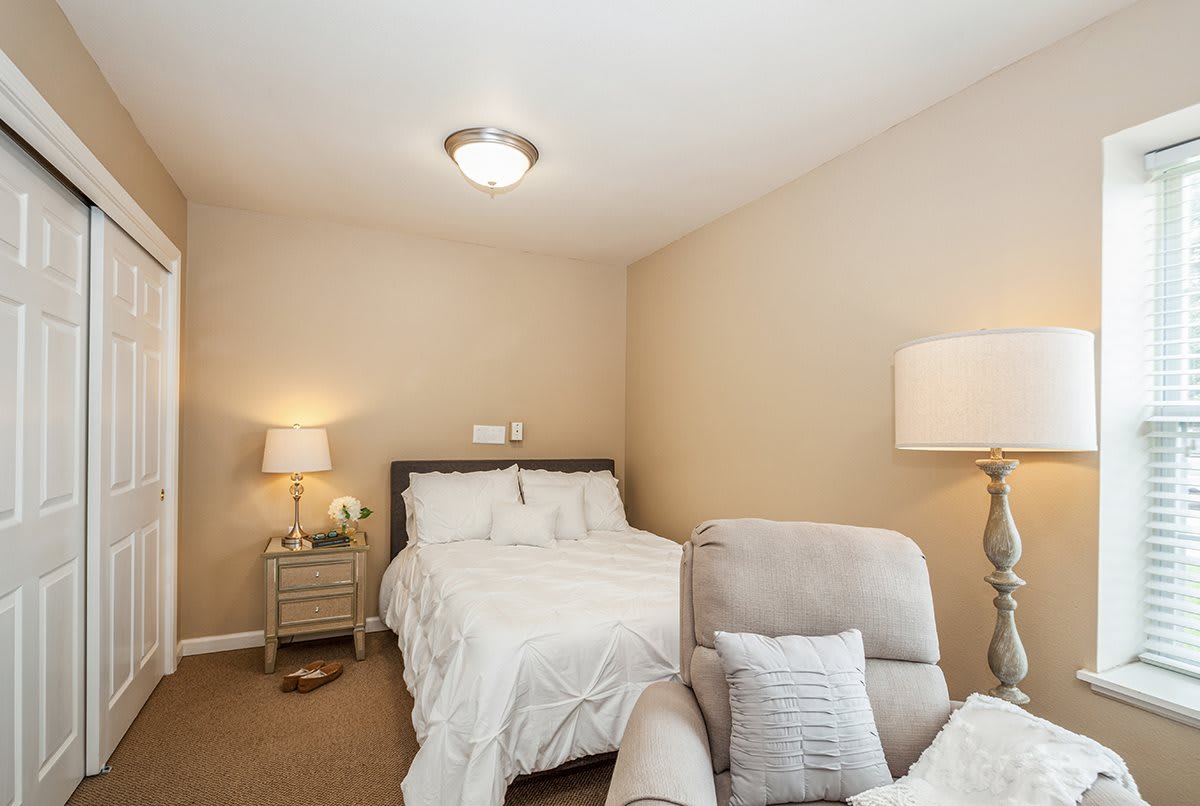 A cozy bedroom featuring a bed with white linens, a nightstand with a lamp, and an armchair in the corner.
