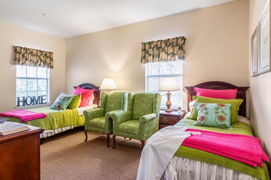 A cozy bedroom with twin beds, vibrant green and pink accents, and comfortable armchairs under natural lighting.