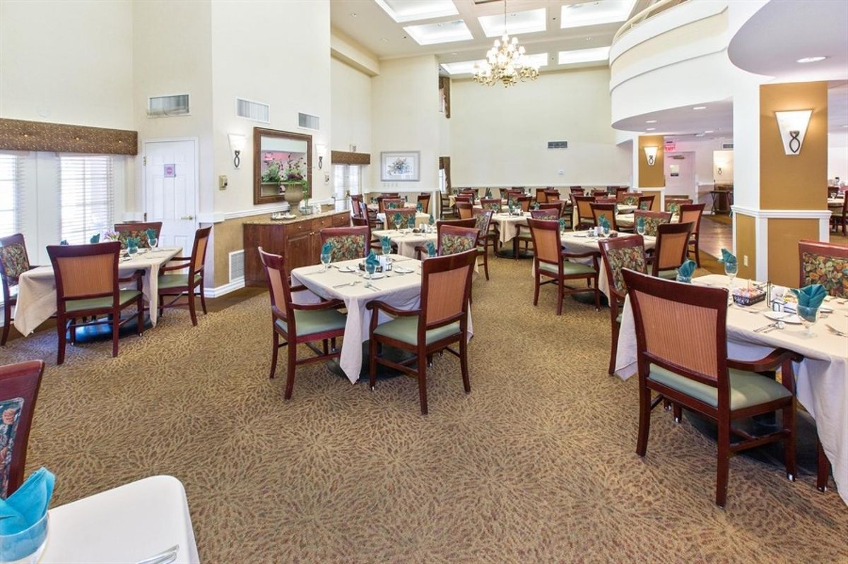A spacious dining room with multiple tables and chairs, featuring a chandelier and carpeted floor.