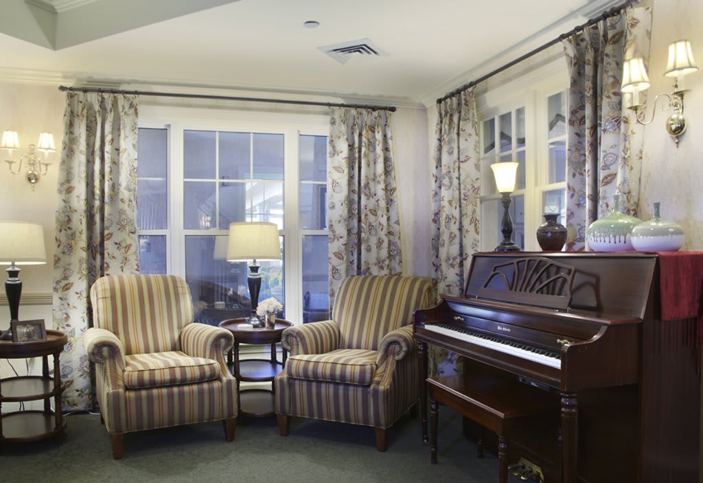 A cozy living room with a piano, two striped armchairs, and a lamp by the window.