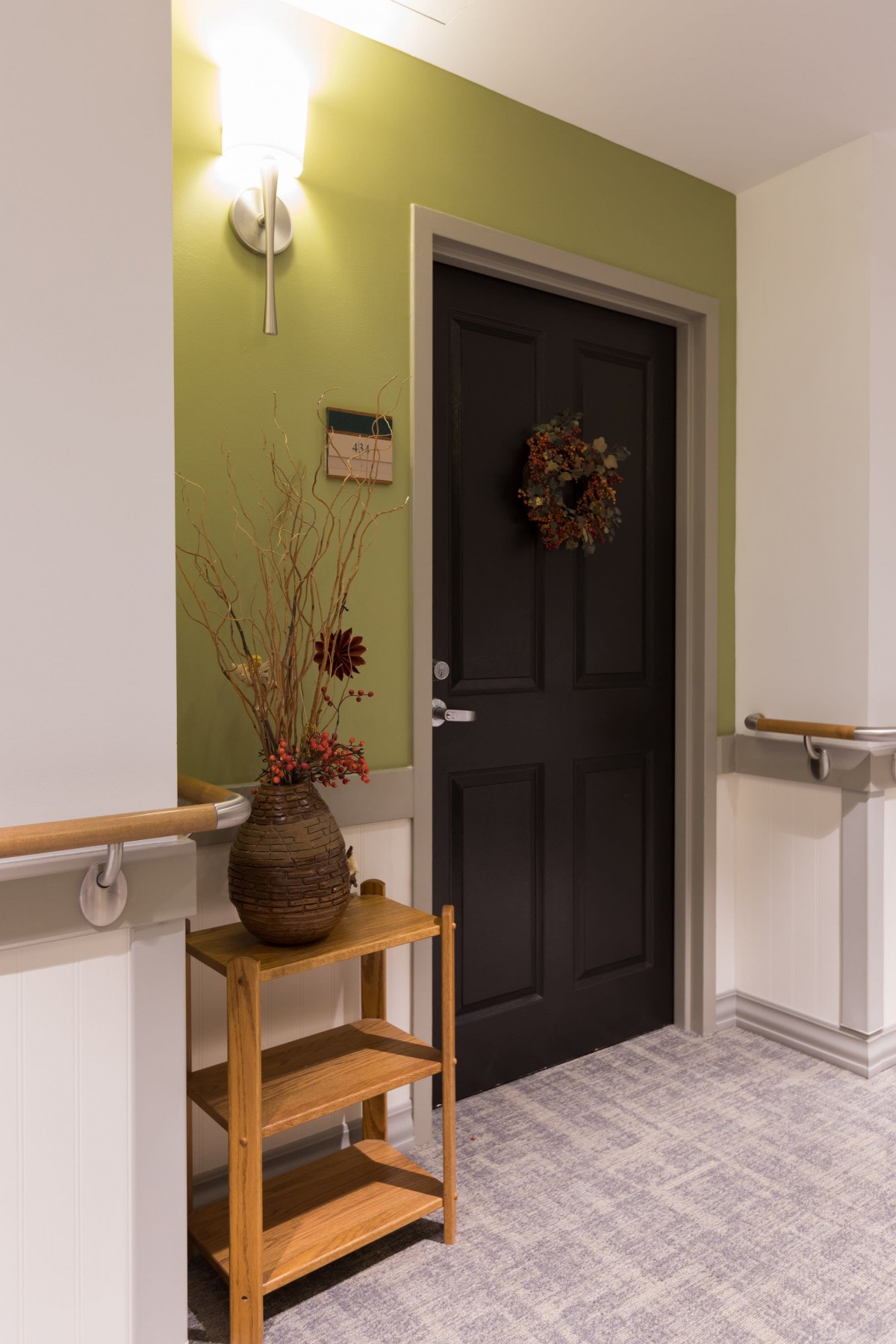 A decorated hallway with a dark door, adorned with a wreath and a vase with dried plants.