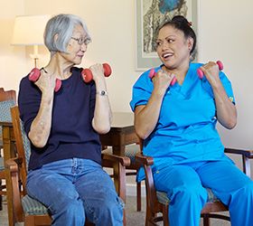An elderly woman and her caregiver perform seated exercises with dumbbells.