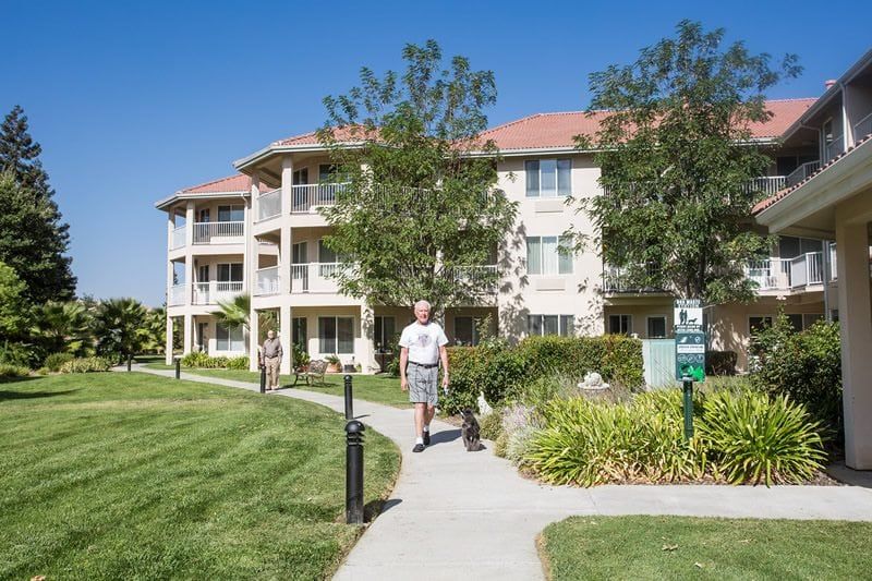 A man walks along a sidewalk in a well-maintained retirement community with landscaped grounds and a multi-story residential building.