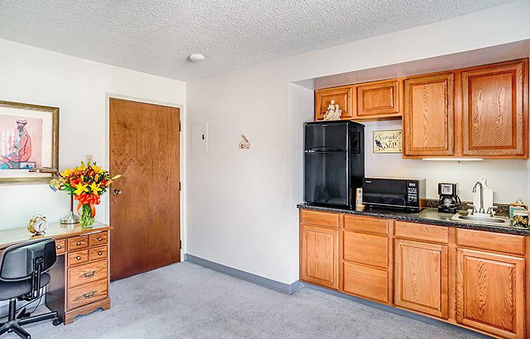 A small kitchenette with wooden cabinetry, featuring a fridge and microwave, and a flower arrangement on a side table.