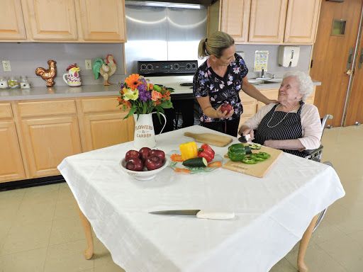 A caregiver assists an elderly person in a kitchen filled with fresh fruits and vegetables.