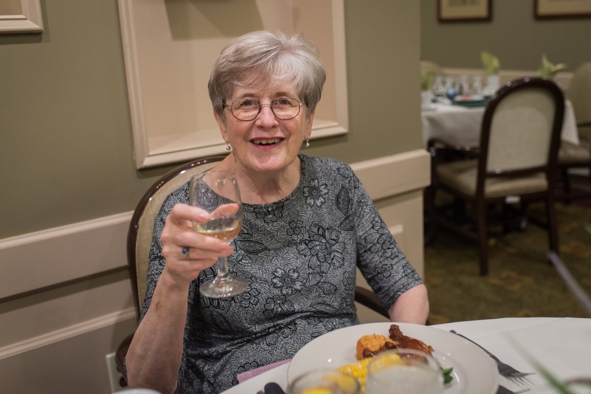 An elderly woman enjoys a meal at a restaurant, raising her wine glass with a smile.