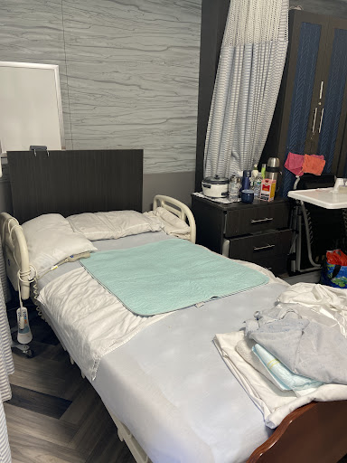 A simple bedroom setting featuring a hospital-style bed with fresh bedding, a nightstand filled with personal items, and draped curtains.