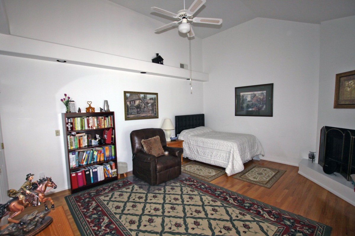 A cozy bedroom featuring a bookshelf, armchair, and single bed, complemented by decorative artwork and a ceiling fan.
