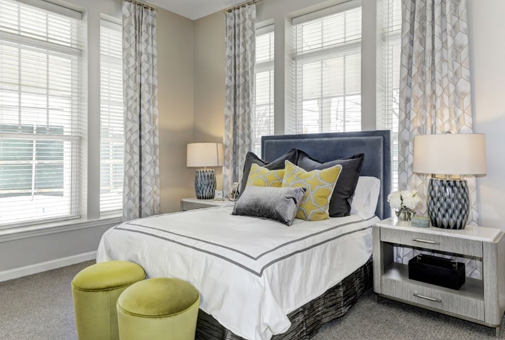 A modern bedroom with stylish decor, featuring a neatly made bed with pillows and two green ottomans at the foot. The room is accented with large windows and curtains.