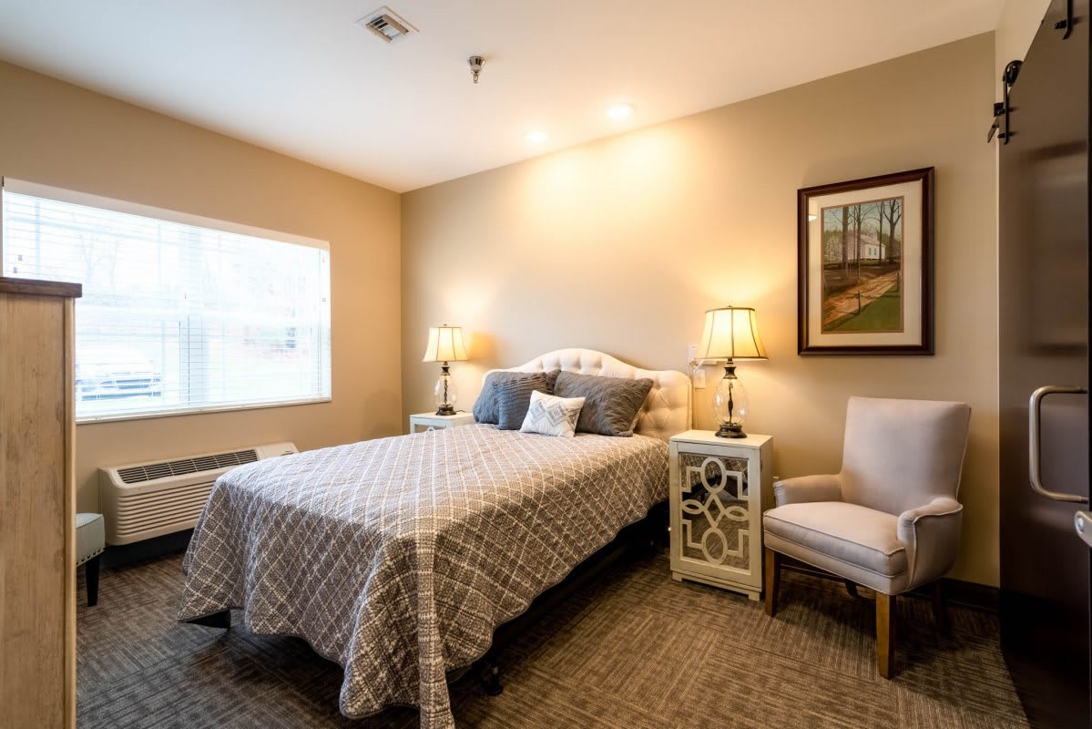 A cozy bedroom with a neatly made bed, two lamps, and a comfortable chair beside a window.