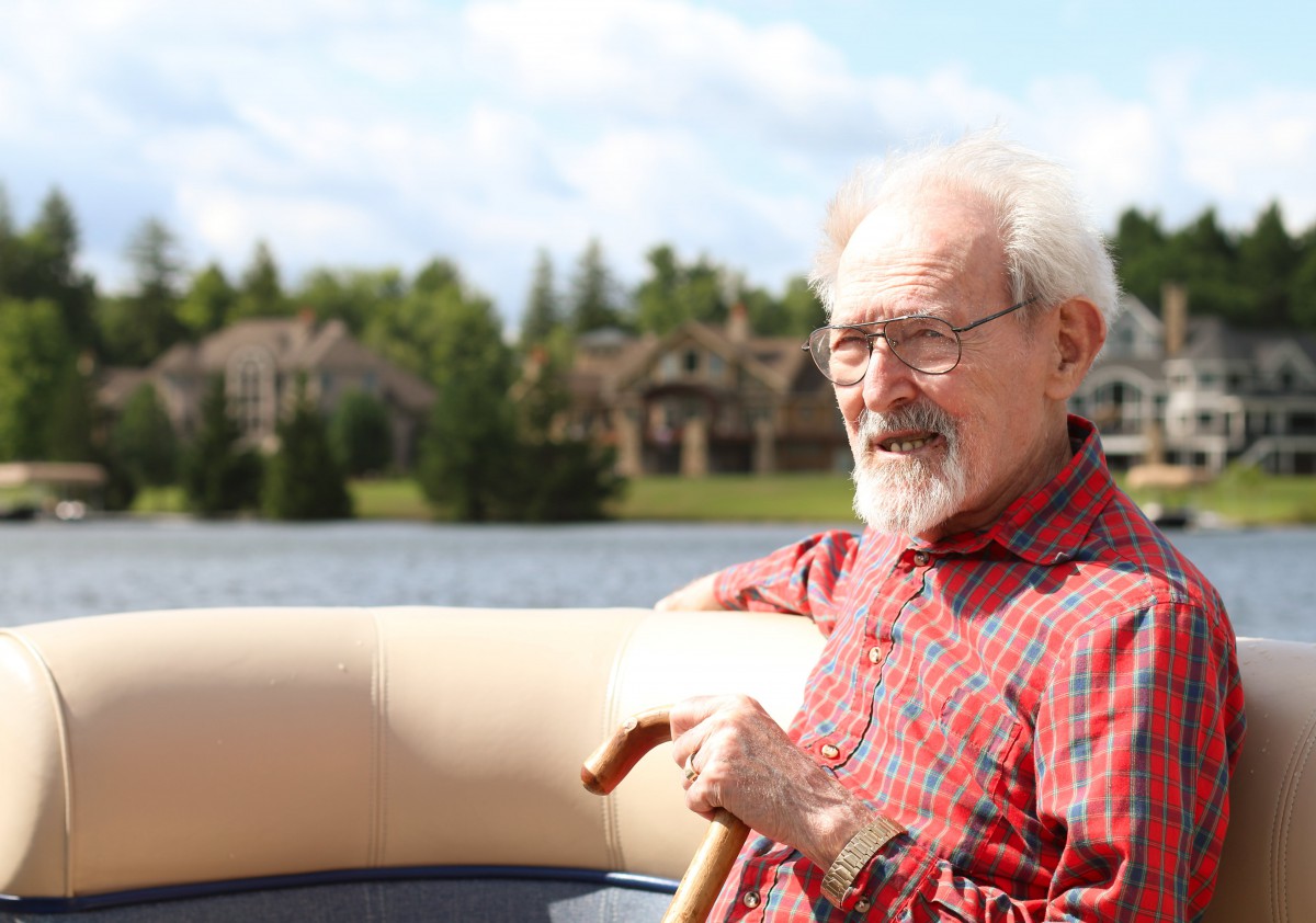 An elderly man sitting on a boat near a lake, holding a cane on a sunny day.