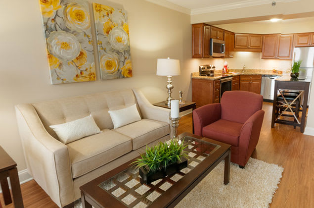 A cozy living room connected to a kitchen, featuring a comfortable sofa, red armchair, decorative artwork, and a glass-top coffee table with a lamp.