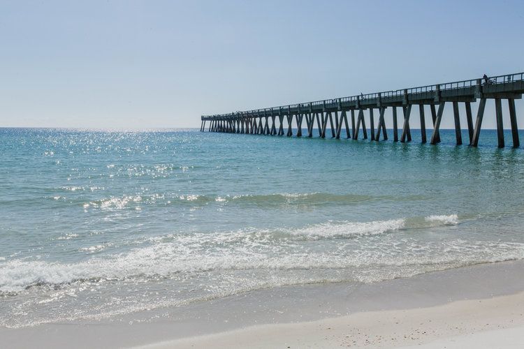 A serene view of a beach with gentle waves and a long pier extending into the ocean under a clear sky.