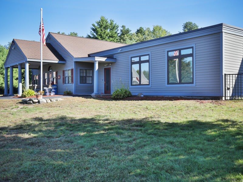 A modern single-story house with a lawn and flagpole, set against a backdrop of trees on a clear day.