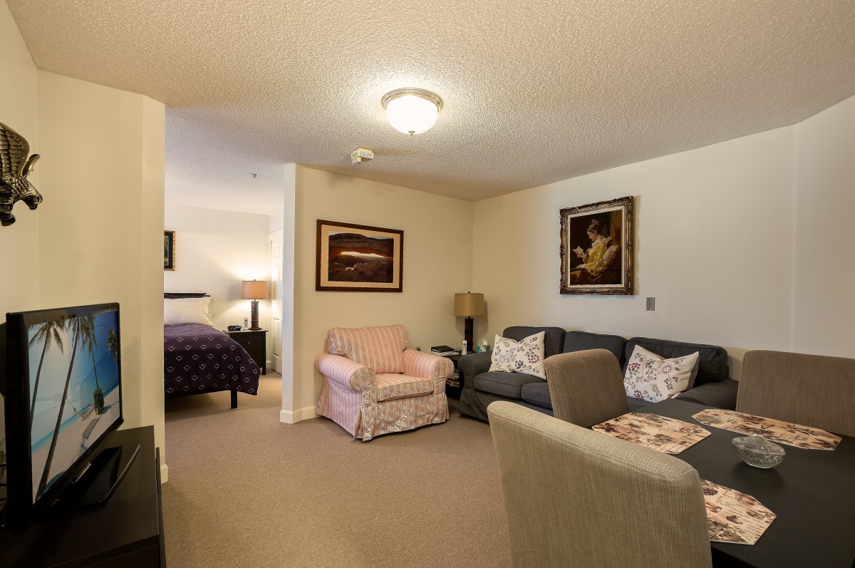 A cozy living room with a view into the bedroom, featuring a sofa, armchair, and decorative paintings on the wall.
