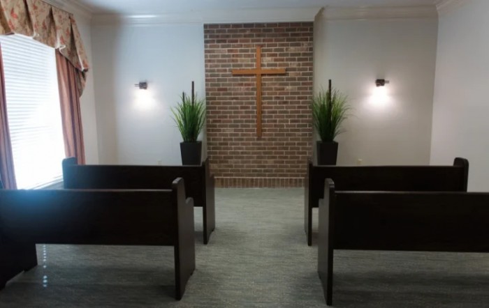 A simple chapel with wooden pews facing a brick wall adorned with a cross, flanked by plants and illuminated by wall lights.