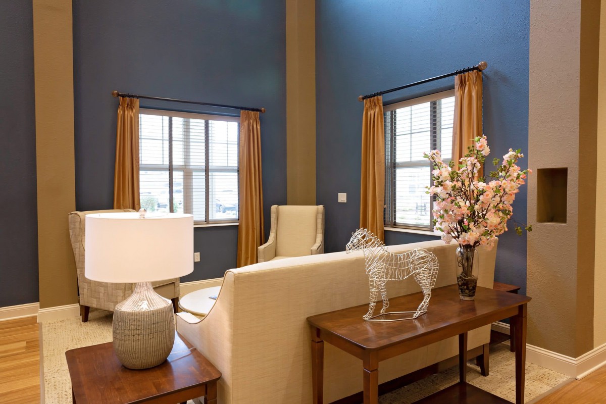 A cozy living room with blue walls, featuring a sofa and armchair, two large windows with golden curtains, and stylish table decor.