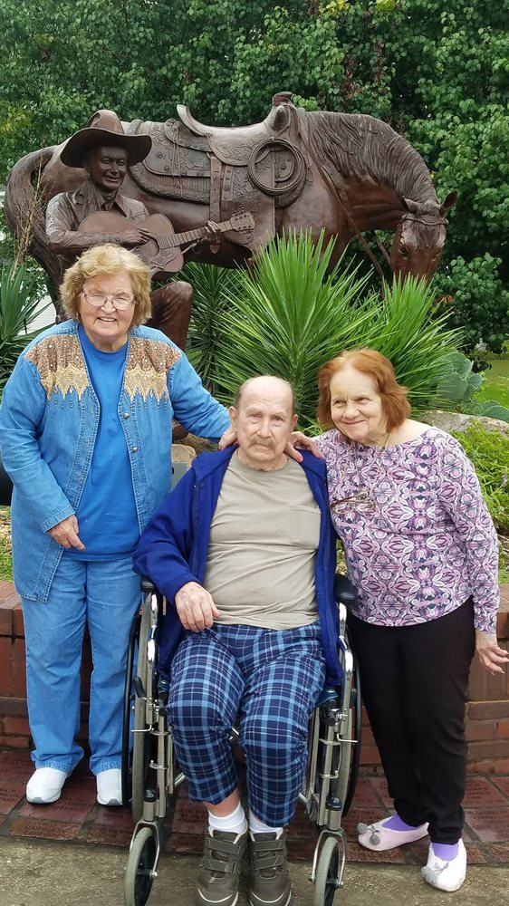 Three elderly individuals, two standing and one seated in a wheelchair, enjoy time together outdoors.