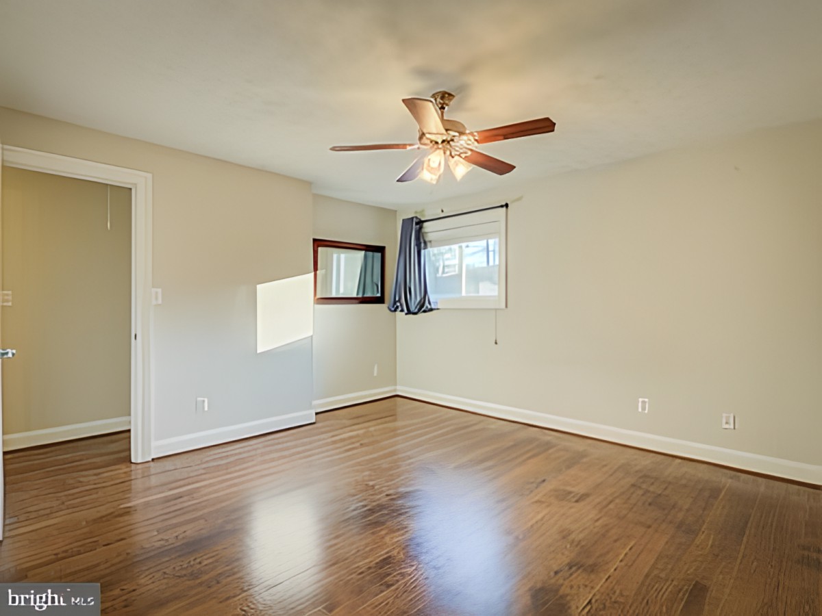 An empty room with wooden flooring and a ceiling fan, featuring large windows that let in natural light.