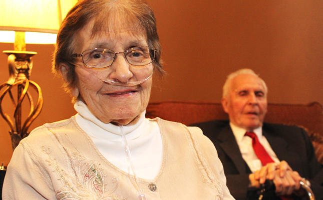 An elderly woman smiling at the camera with a man in formal attire sitting in the background.