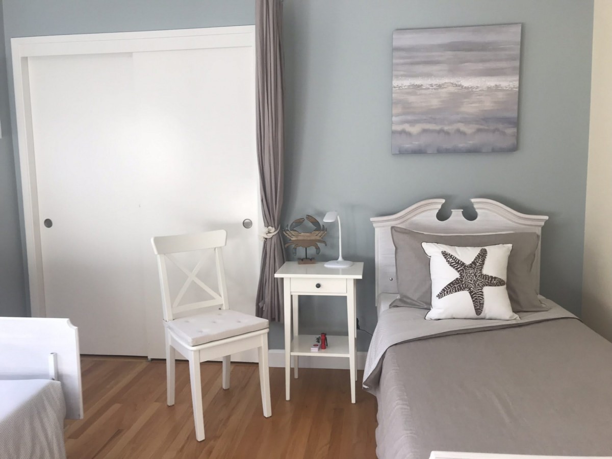 A cozy coastal-themed bedroom with neutral tones, featuring a bed with a starfish pillow, a white chair, and a small nightstand.