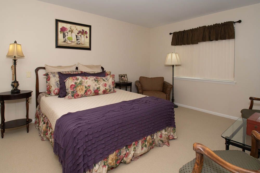 A cozy bedroom with floral-patterned bedding and a purple throw blanket, featuring a lamp and a comfortable chair in the corner.