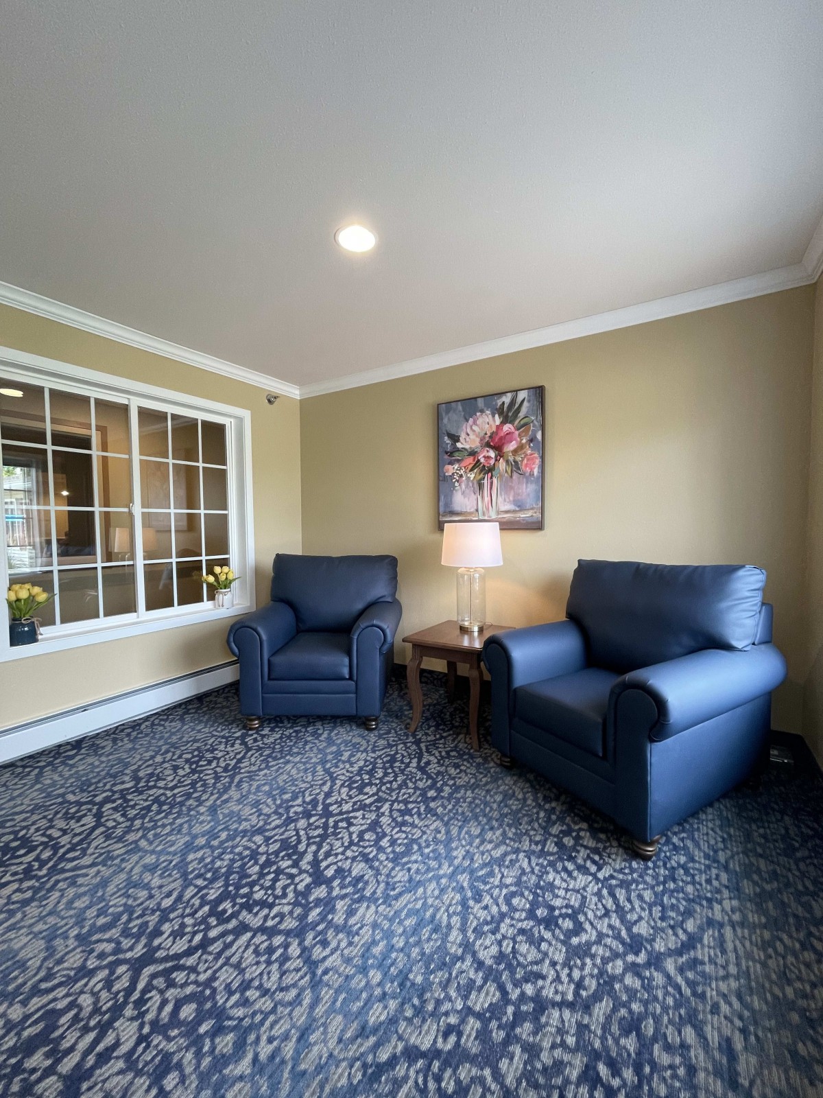 A cozy living room with two blue armchairs, a table with a lamp, and a decorative painting on the wall.