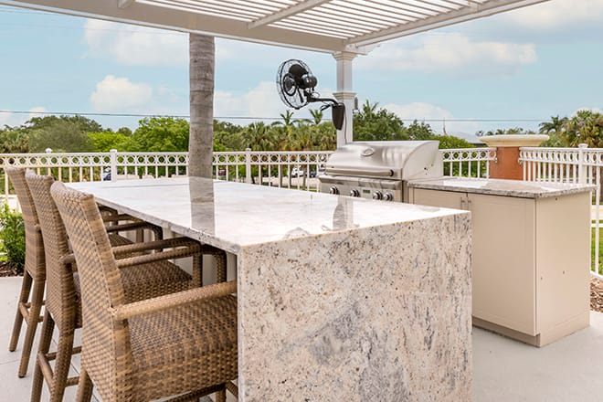 A modern outdoor kitchen with a marble countertop, barbecue grill, and wicker seating, set under a pergola with a scenic nature view.