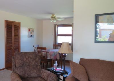 A cozy living room with a view of the adjoining dining area, featuring comfortable sofas, a table lamp, and a window bringing in natural light.