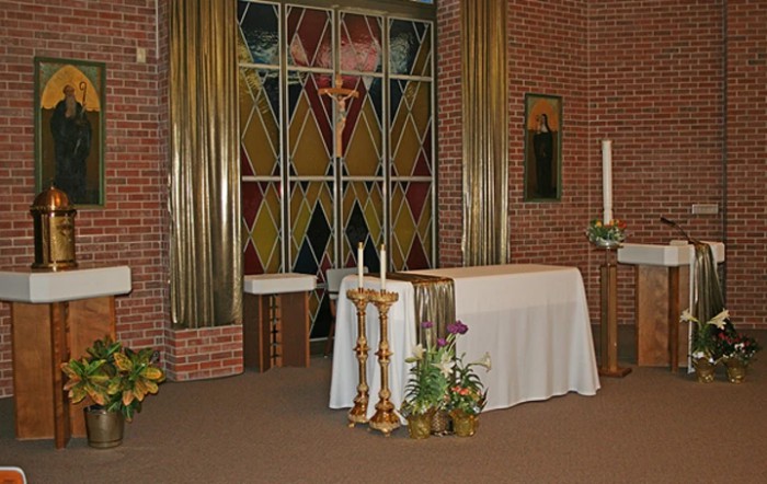 A church interior featuring an altar with flower arrangements and a backdrop of stained glass windows.