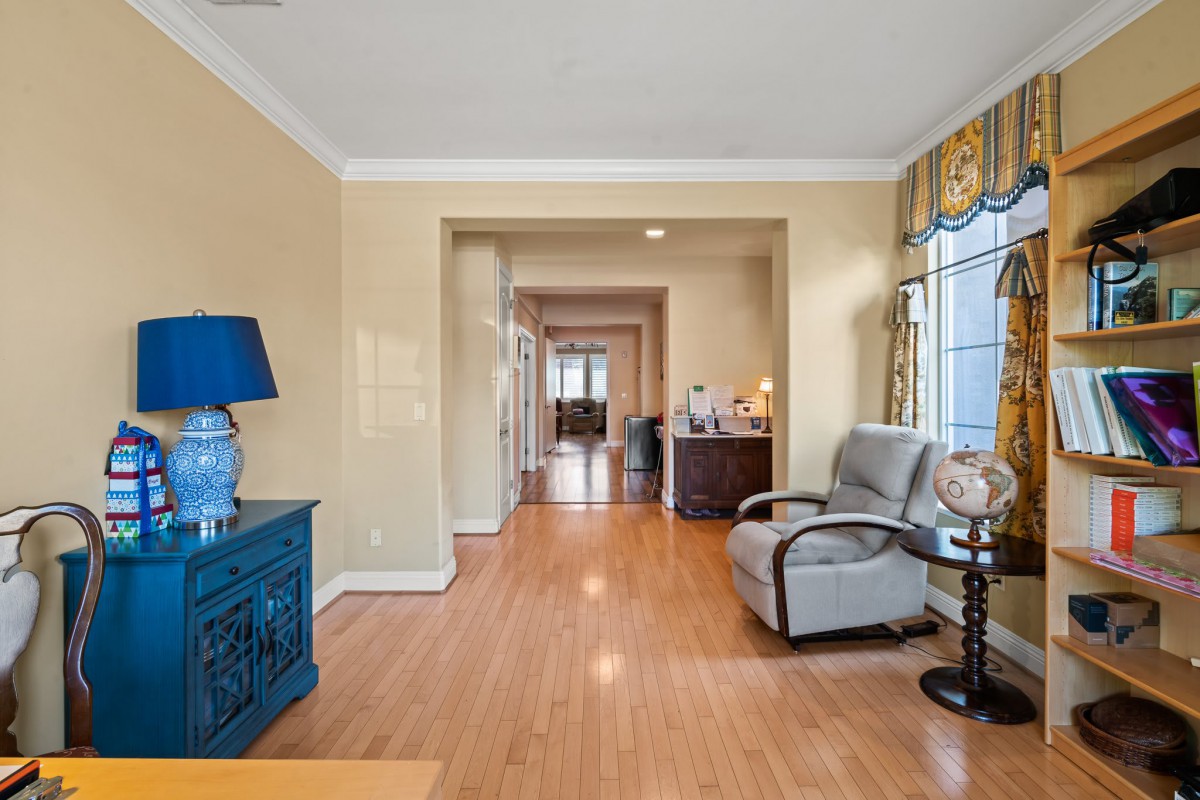 A cozy living room with hardwood flooring and a view into the hallway, featuring an armchair, a lamp on a blue dresser, and natural light streaming through the window.