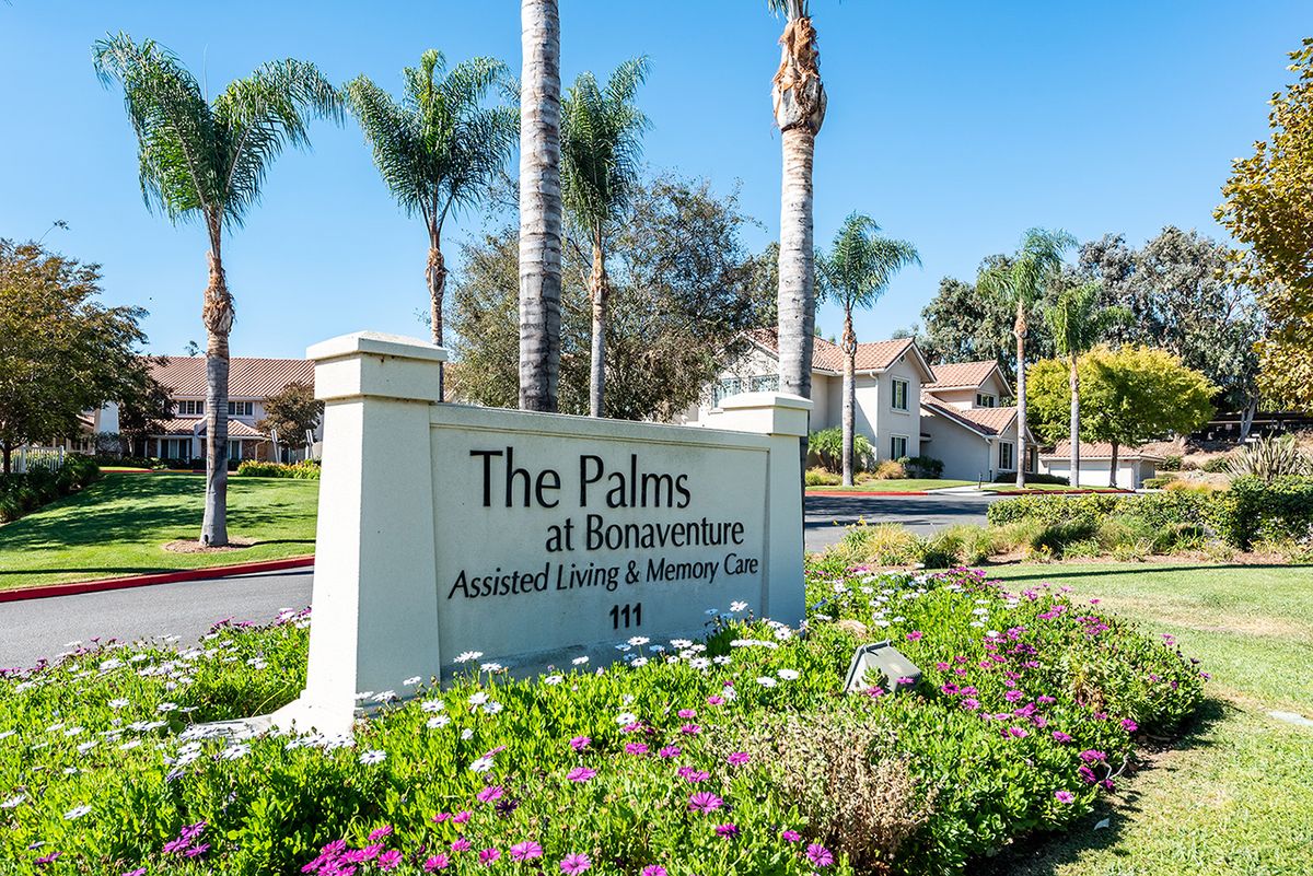 The entrance sign of The Palms at Bonaventure, an assisted living and memory care community, surrounded by palm trees and colorful flowers.