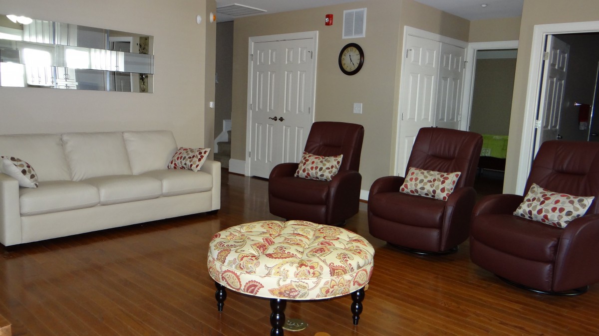 A cozy living room featuring a white sofa, two brown recliners with patterned pillows, and a floral ottoman on a hardwood floor.