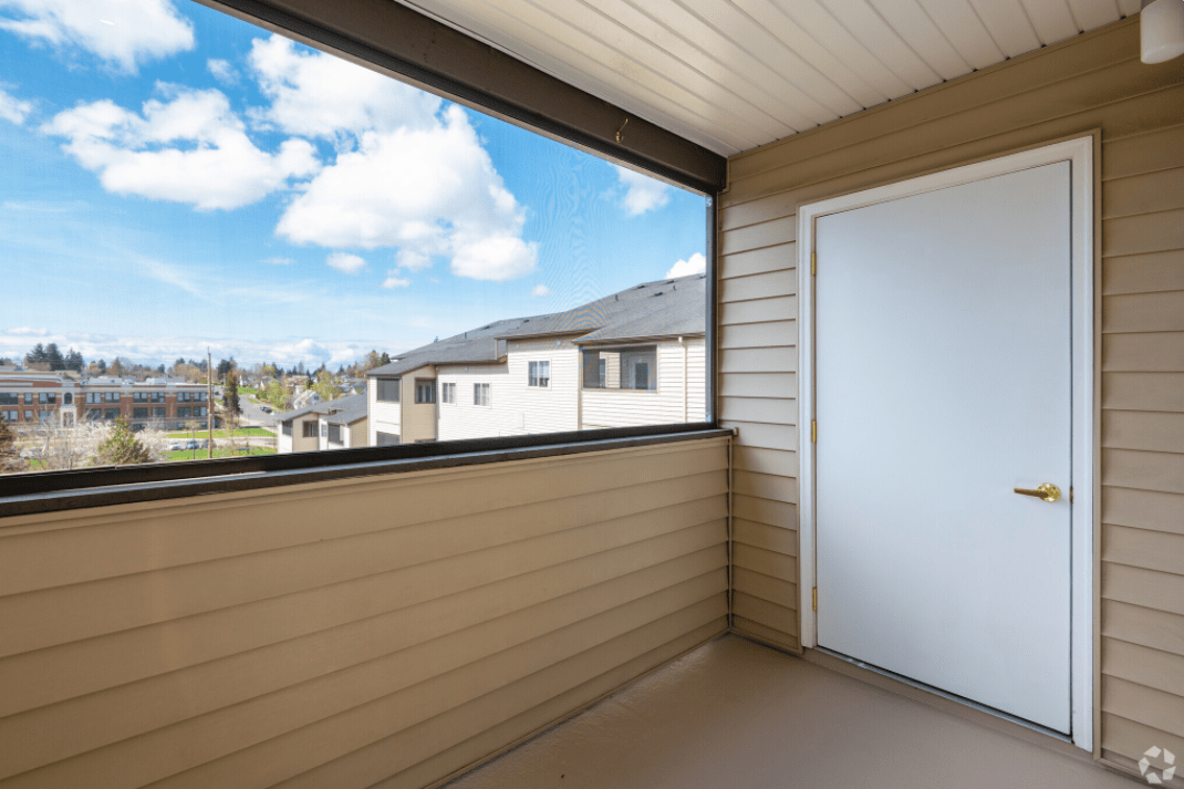 A residential balcony overlooking a neighboring building, featuring a clear blue sky and clouds.