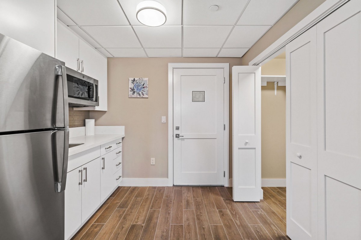A modern kitchen area with white cabinets and a microwave, featuring wooden flooring and a door leading to another room.