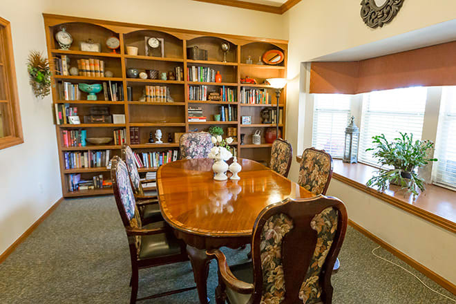 A cozy library room with a wooden table surrounded by ornate chairs and large bookshelves filled with books.