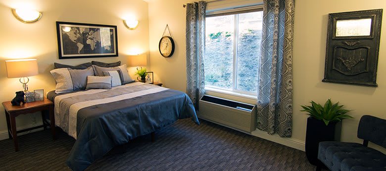 A cozy bedroom with a neatly made bed, wall-mounted clock, and large window letting in natural light.