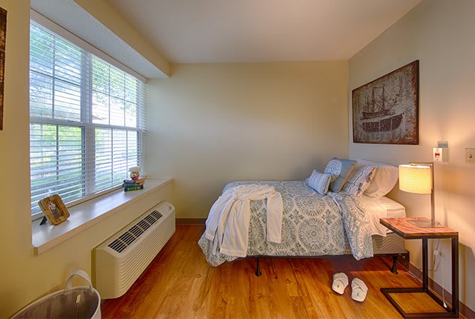 A cozy bedroom with a neatly made bed, nightstand, and large window letting in natural light.