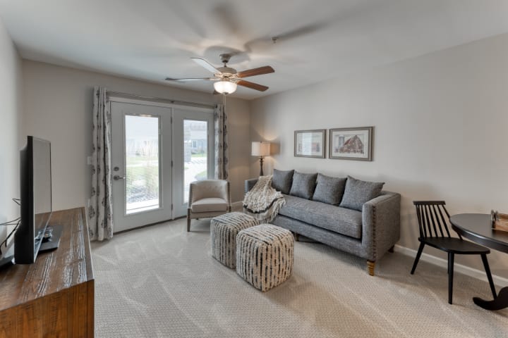 A cozy living room with neutral tones, featuring a sofa, armchair, and ottomans under a ceiling fan.