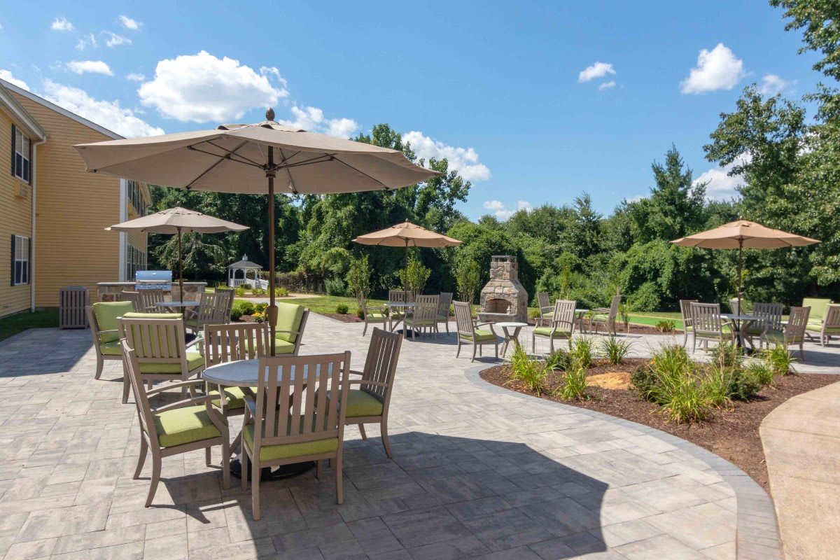 A spacious outdoor patio with tables, chairs, and umbrellas set against a lush garden backdrop on a sunny day.